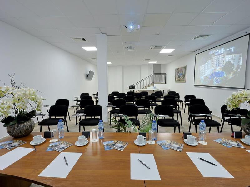 Vergaderruimte met een lange houten tafel vooraan, stoelen en een projectiescherm.