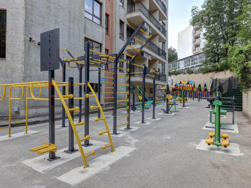 Buitenfitnessruimte met trainingsapparatuur naast een woongebouw onder bomen.