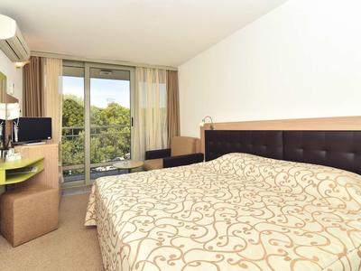 Lichte hotelkamer met tweepersoonsbed, grote ramen en balkon met uitzicht op groen.