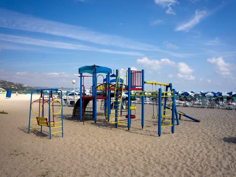 Kleurrijke speeltuin met schommels en klimrek op zandstrand onder blauwe lucht.