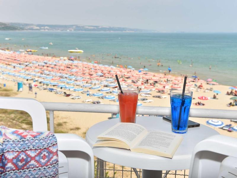 Uitzicht op een druk zandstrand met parasols, een tafel met twee drankjes en een boek op de voorgrond.