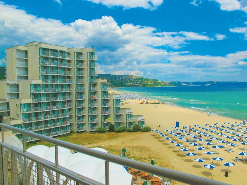 Hotel met moderne glazen gevel naast een zandstrand met parasols en blauwe lucht.