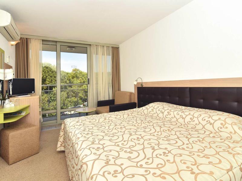 Lichte hotelkamer met tweepersoonsbed, grote ramen en balkon met uitzicht op groen.