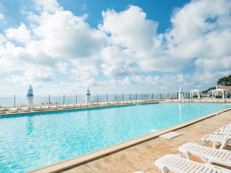 Großer Außenpool mit Liegestühlen unter bewölktem Himmel neben einem Hotel.