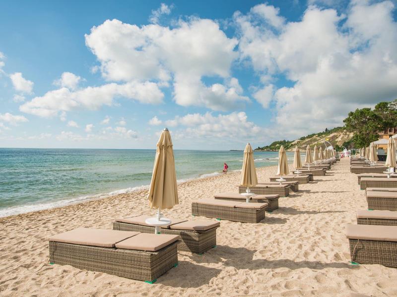 Strand mit Liegestühlen und geschlossenen Sonnenschirmen unter blauem Himmel mit Wolken.