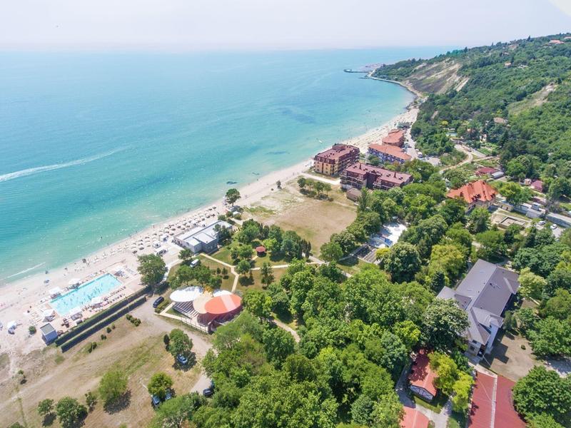 Luftaufnahme eines Strandes mit türkisfarbenem Wasser, bewaldeter Küste und Gebäuden am Ufer.