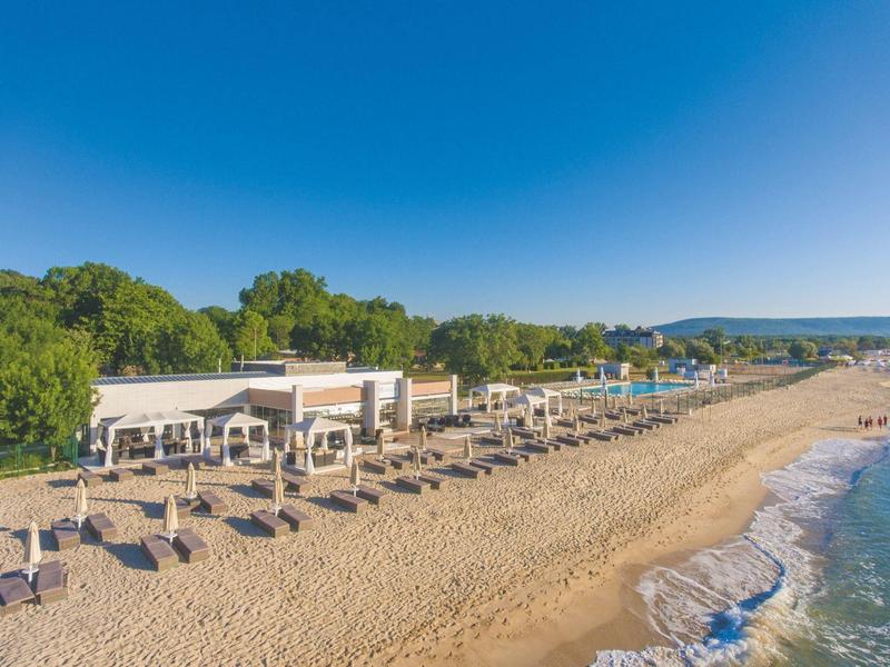 Langer Sandstrand mit Liegen und Sonnenschirmen neben einem Hotelgebäude unter klarem blauem Himmel.