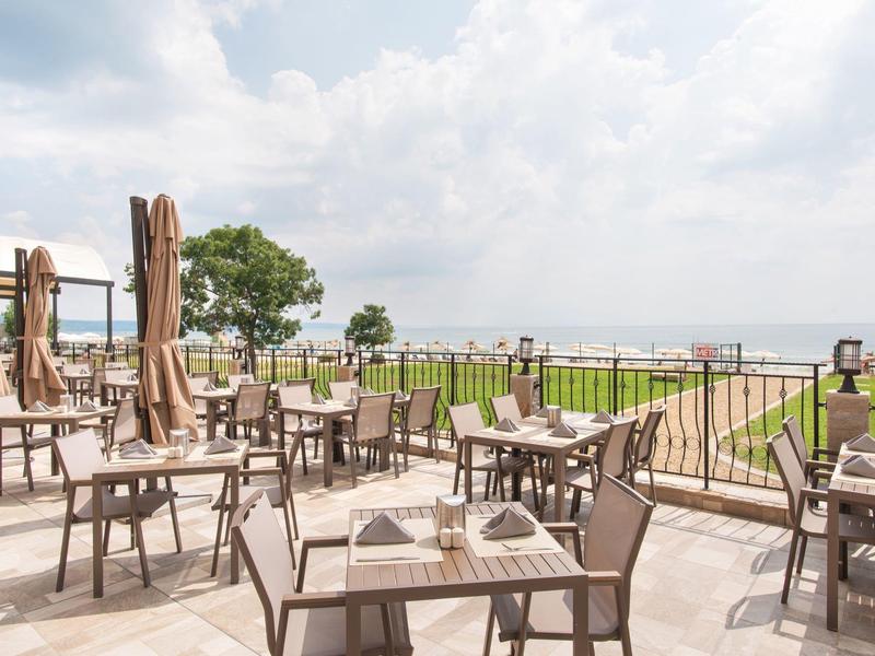 Leere Außenterrasse mit Tischen, Stühlen und Sonnenschirmen mit Blick auf den Garten und Wasser.
