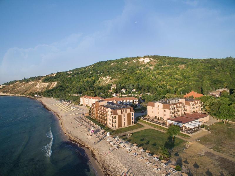 Luftaufnahme eines Strandes mit Hotels vor einem bewaldeten Hügel unter blauem Himmel.