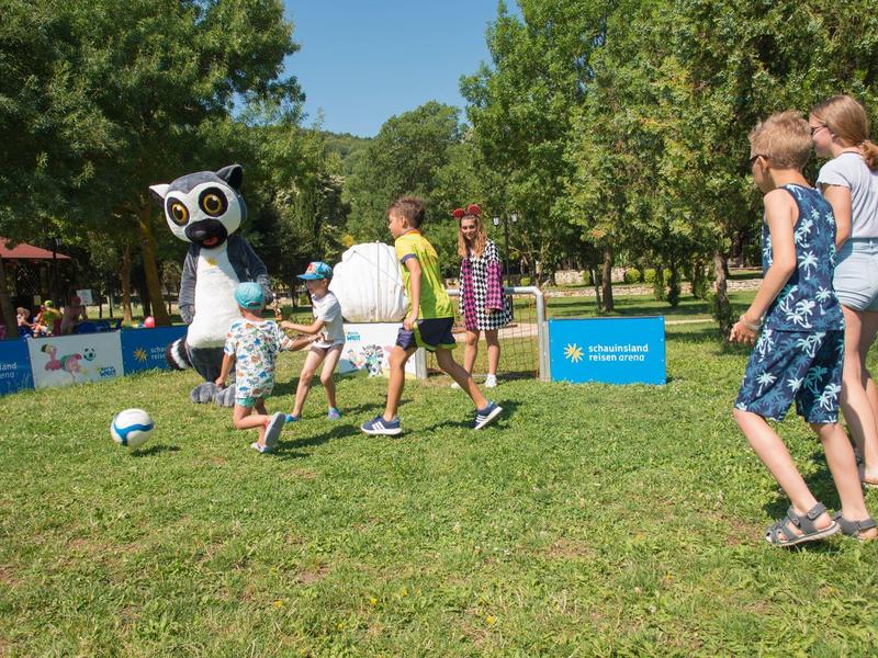 Kinder spielen Fußball mit einer Robotfigur im Park bei sonnigem Wetter.