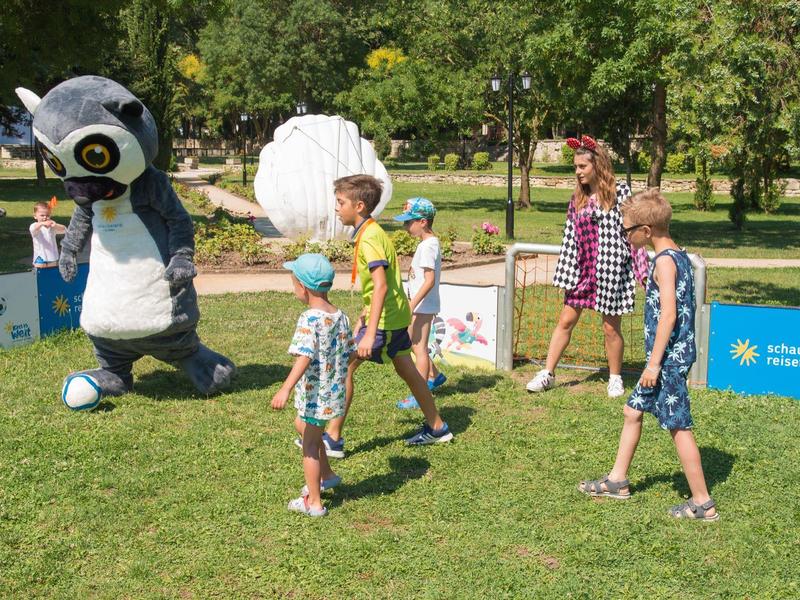 Kinder laufen auf einer Wiese in einem Park, begleitet von einer Person in einem lustigen Panda-Kostüm.