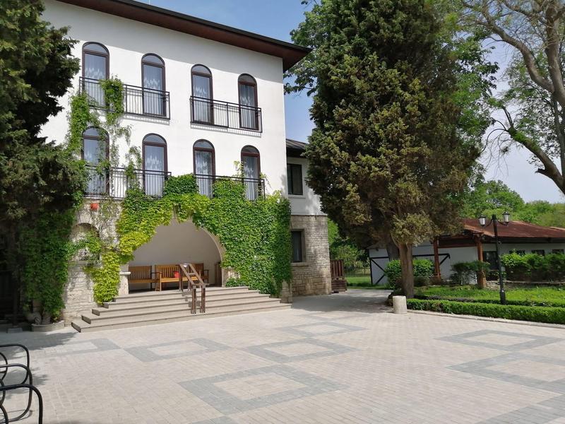 Mehrstöckiges Hotel mit weißen Wänden, Rundbogenfenstern und grüner Kletterpflanze, umgeben von Bäumen.