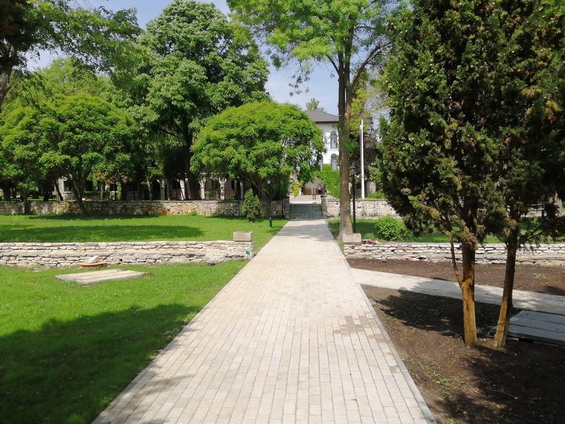 Gerader gepflasterter Weg führt durch einen grünen Park mit Bäumen und einer historischen Mauer.