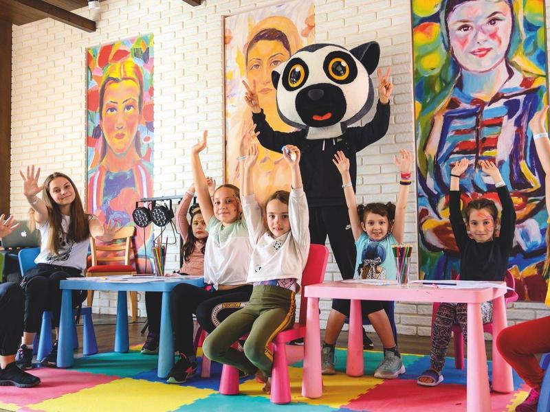 Kinder sitzen an bunten Tischen und machen fröhlich gemeinsam den Victory-Gruß in einem Raum mit farbenfrohen Wandbildern.