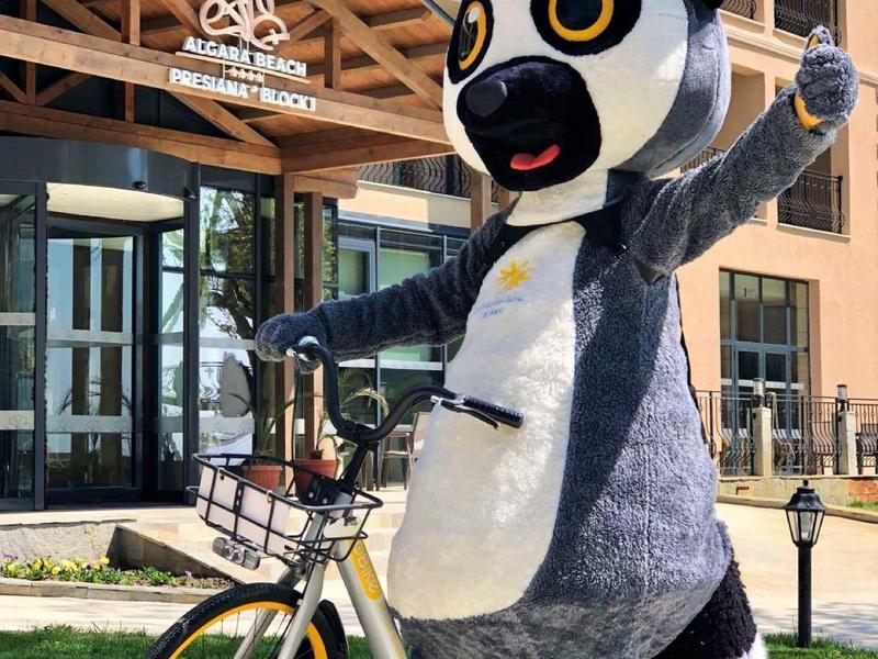 Große Pandafigur mit gelben Augen auf Fahrrad vor modernem Gebäude mit Holzverkleidung.