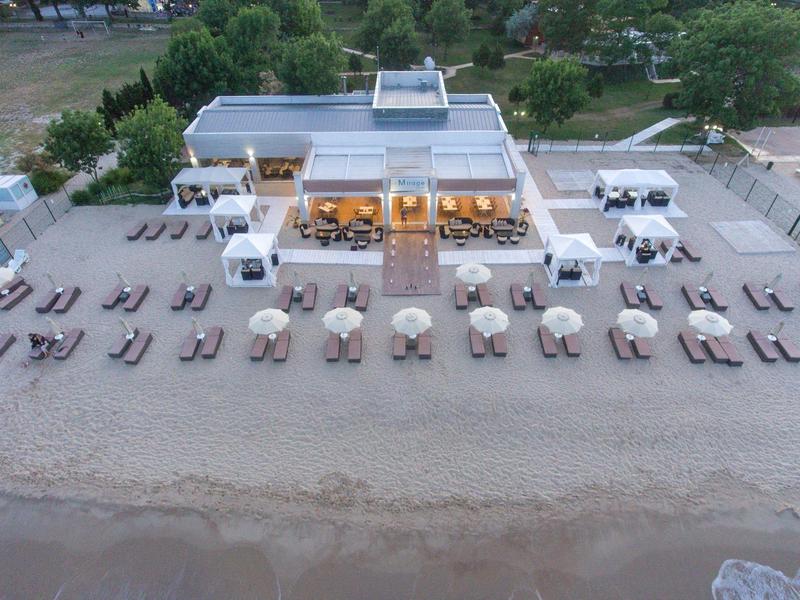 Strandhotel mit Liegestühlen, Sonnenschirmen und einem offenen Restaurantbereich direkt am Sandstrand.