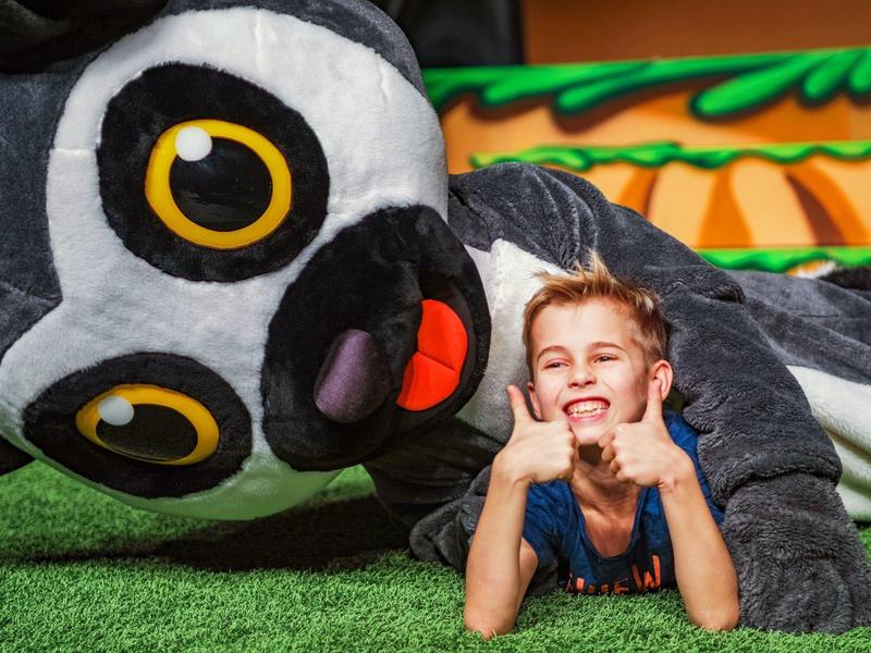 Kind liegt lachend auf Gras neben großem Lemuren-Kostüm im Freien