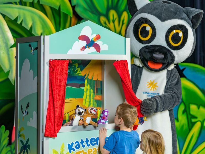 Kinder sehen ein Marionettentheater mit roten Vorhängen und einem Kuscheltier auf der Bühne.