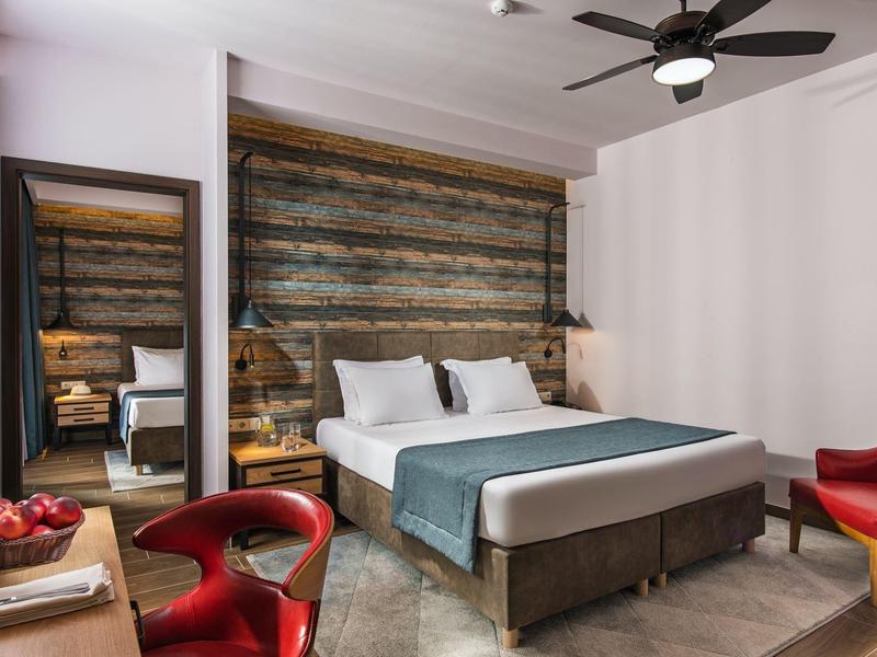 Modernes Hotelzimmer mit Doppelbett, Holzakzentwand und roten Stühlen neben einem Spiegel.