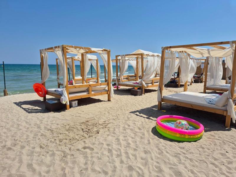 Vier hölzerne Baldachin-Liegen am Strand mit Blick auf das Meer und einen bunten Kinderpool.