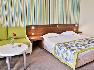 Lichte hotelkamer met tweepersoonsbed, groene bank en ronde tafel met bloem.