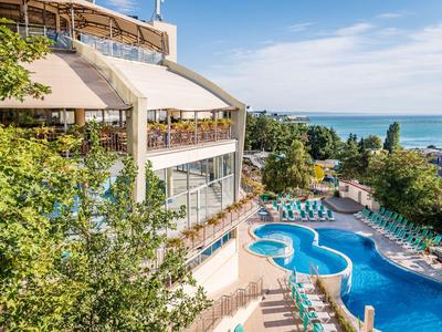 Widok na hotel z zakrzywionym basenem i leżakami, otoczony drzewami z widokiem na morze.