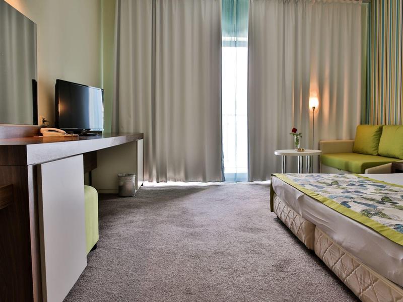 Moderne hotelkamer met groot bed, tv, bureau en groene bank in warm licht.