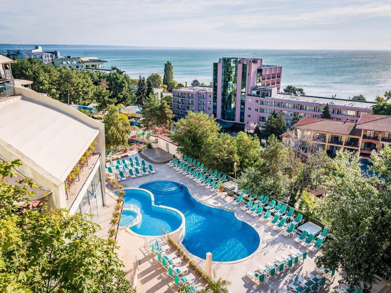 Hotelzwembad met ligstoelen omgeven door bomen, uitzicht op zee en meerdere gebouwen.