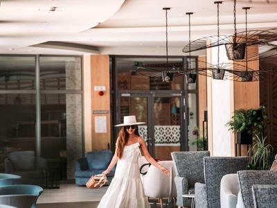 Žena v bílých šatech a klobouku kráčí v elegantní hotelové restauraci s moderním zařízením.