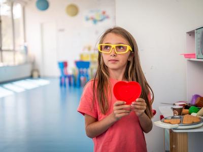 Dívka s žlutými brýlemi drží červené srdce v světlé moderní místnosti.