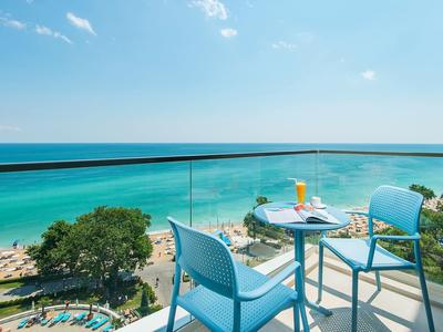 Výhled z balkonu se skleněným zábradlím na pláž a tyrkysové moře pod modrou oblohou.