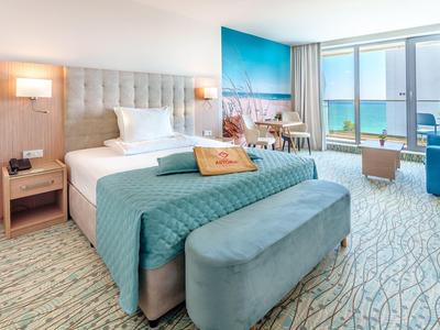 Moderní hotelový pokoj s velkou postelí, modrým dekorem a výhledem na moře přes velká okna.