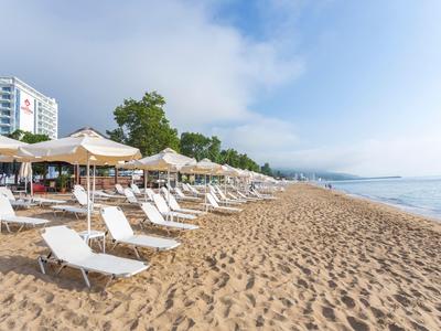 Písčitá pláž s lehátky a slunečníky vedle hotelové budovy za jasné oblohy.