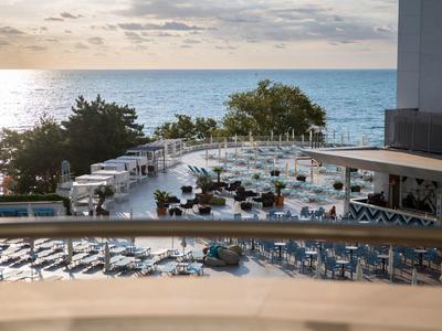 Pohled na hotelový bazén s lehátky a stoly u moře při západu slunce.