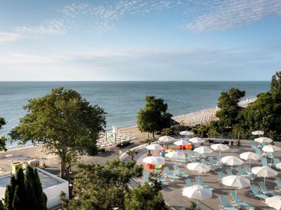 Vue sur une piscine avec des parasols et des arbres au bord de la mer sous un ciel bleu.
