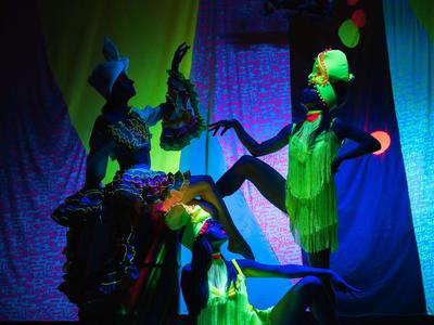 Trois personnes vêtues de couleurs vives dansent devant un décor coloré sous un éclairage théâtral.