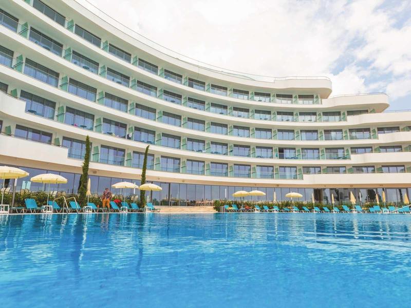 Modern hotelcomplex met groot buitenzwembad en parasols onder een bewolkte lucht.