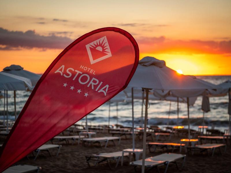 Červený banner Astoria před pláží s západem slunce a zavřenými bílými slunečníky.