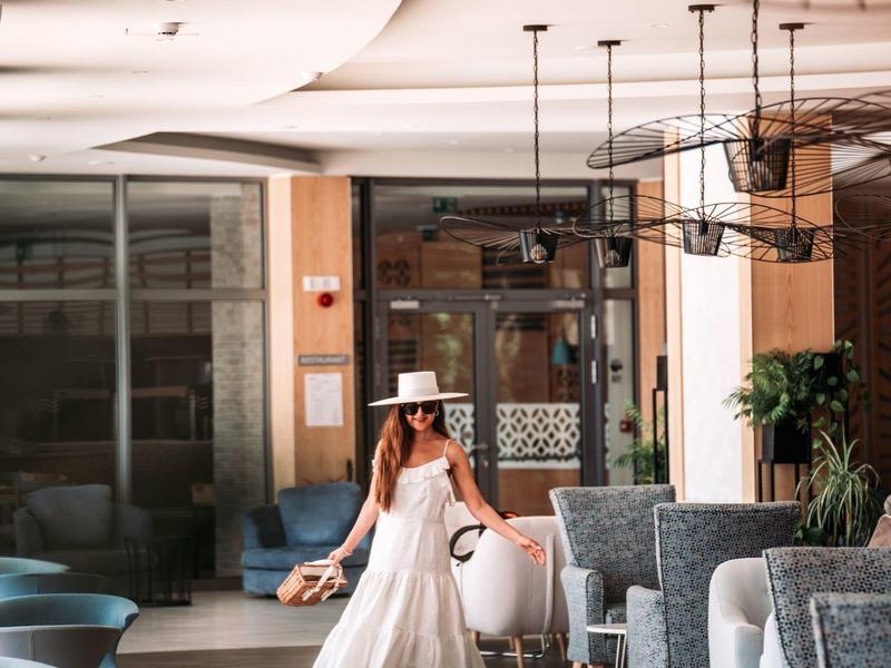 Žena v bílých šatech a klobouku kráčí v elegantní hotelové restauraci s moderním zařízením.