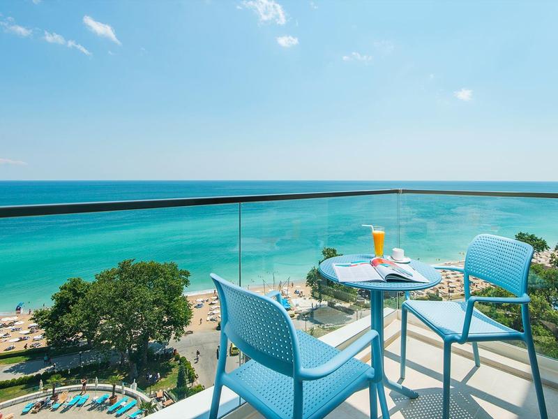 Výhled z balkonu se skleněným zábradlím na pláž a tyrkysové moře pod modrou oblohou.