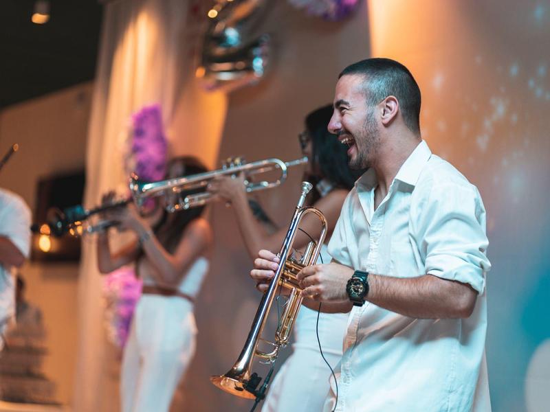 Muž hraje na trubku při slavnostní události s dalšími hudebníky v pozadí.