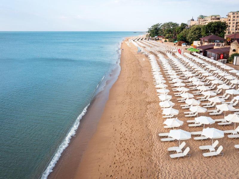 Široká písečná pláž s řadami bílých lehátek a slunečníky vedle klidného moře.