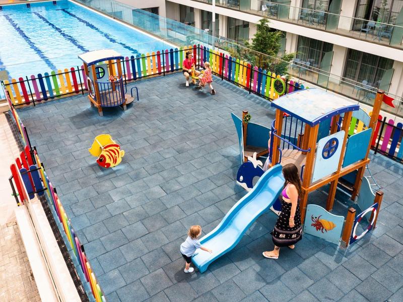 Dětské hřiště s barevným plotem vedle hotelového bazénu.