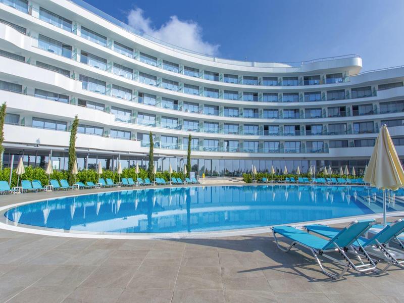 Moderní hotel s zakřivenou budovou a bazénovou částí s lehátky a slunečníky.