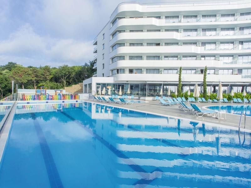 Velký hotelový bazén s slunečníky a lehátky před moderní bílou hotelovou budovou.