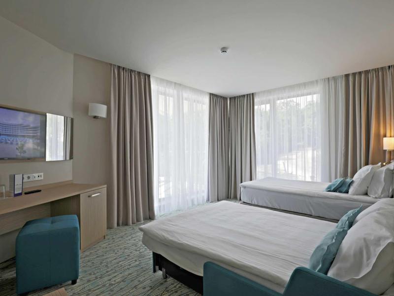 Modern hotelkamer met twee bedden, bureau, tv en grote ramen met gordijnen.