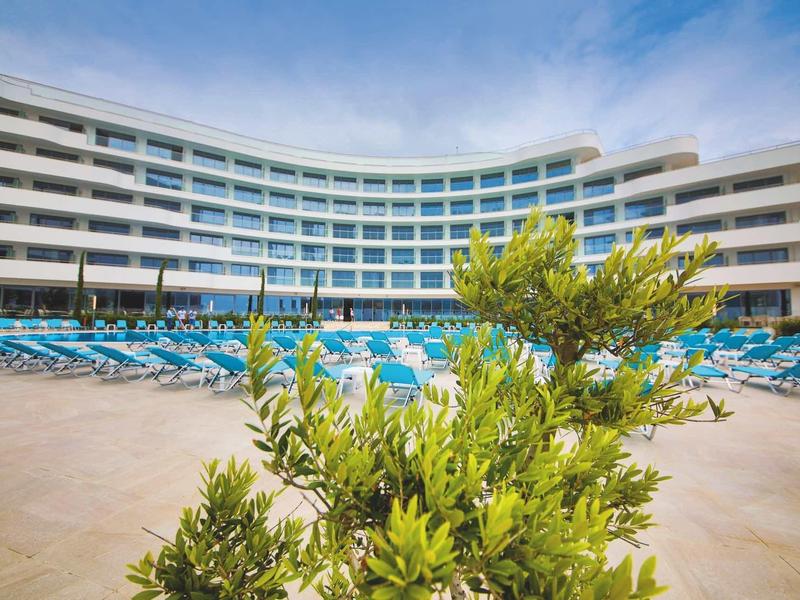 Groot hotel met gebogen gevel en lege blauwe ligstoelen bij het zwembad.