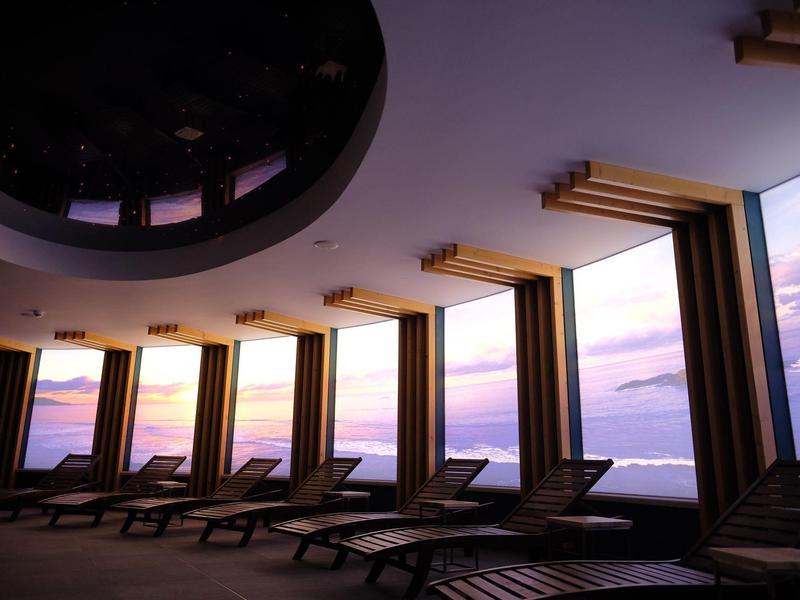 Řady lehátek vedle velkých oken s výhledem na západ slunce nad mořem.