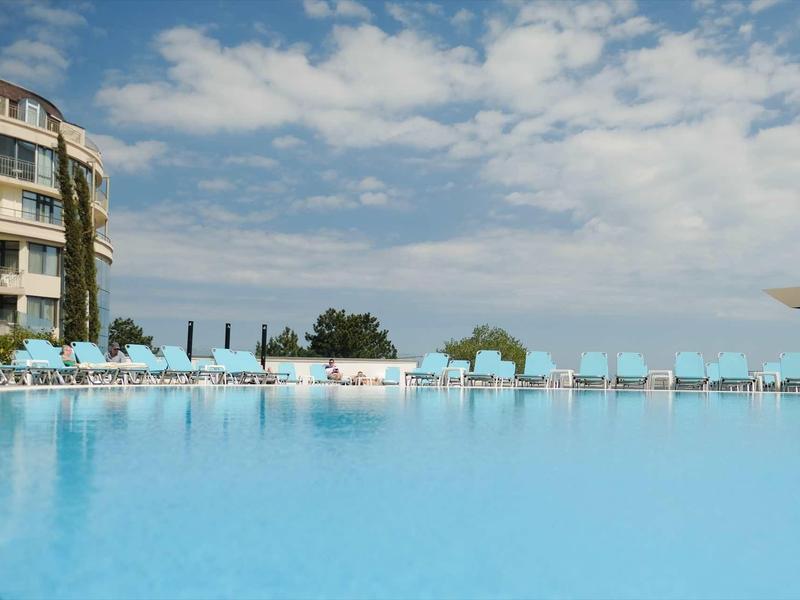 Groot hotelzwembad met ligstoelen voor een meerlaagse gebouw onder een blauwe lucht.