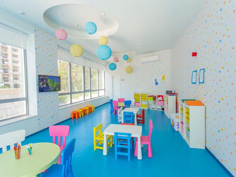 Barevná dětská herna se stoly, židlemi a oknovou zdí v jasné místnosti.